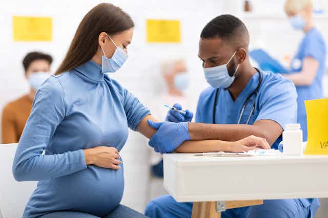 A pregnant white woman wearing a face mask with her hand on her belly receives an injection from a Black health-care professional in scrubs and a face mask.