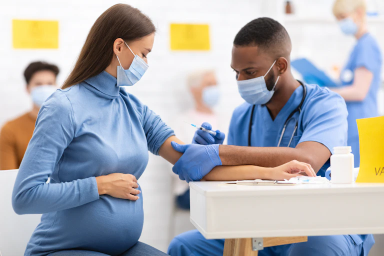 A pregnant white woman wearing a face mask with her hand on her belly receives an injection from a Black health-care professional in scrubs and a face mask.
