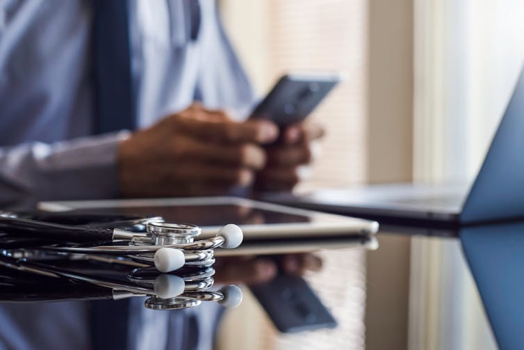 A doctor uses a smartphone. His stethoscope, tablet and laptop sit on the table.