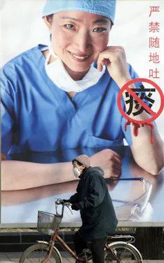 A woman on a bicycle and wearing a mask rides past a billboard showing a picture of a health worker.