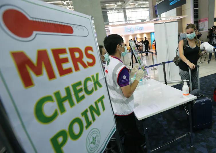 A woman with a mask and a suitcase stands at MERS checkpoint staffed by a healthworker at an airport
