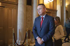 A man wearing a blue suit and a red tie stands in a government building with his hands clasped in front of him.