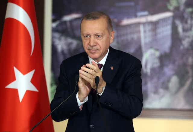 Turkey's Recep Tayyip Erdogan applauds as he stands behind a microphone and beside the Turkish flag. 