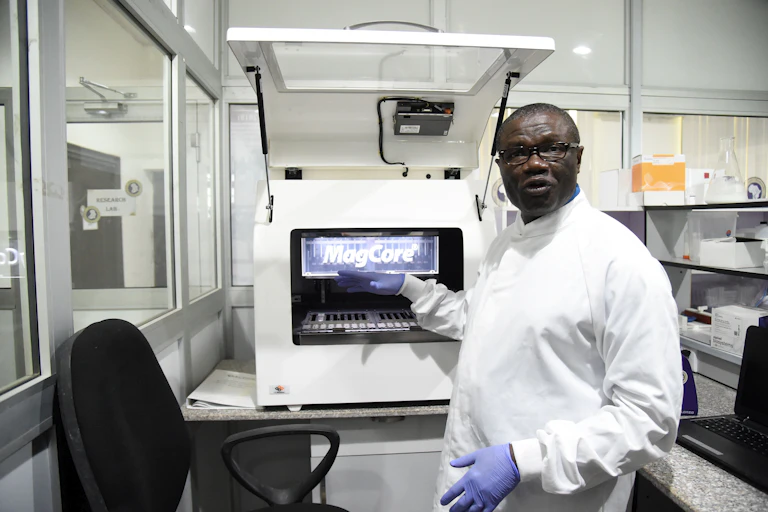 A man in a white overall lab coat points at a machine in the laboratory.