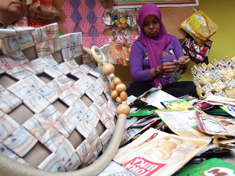 Seorang perempuan sedang memilin kantong plastik dikelilingi tas daur ulang dari kemasan.