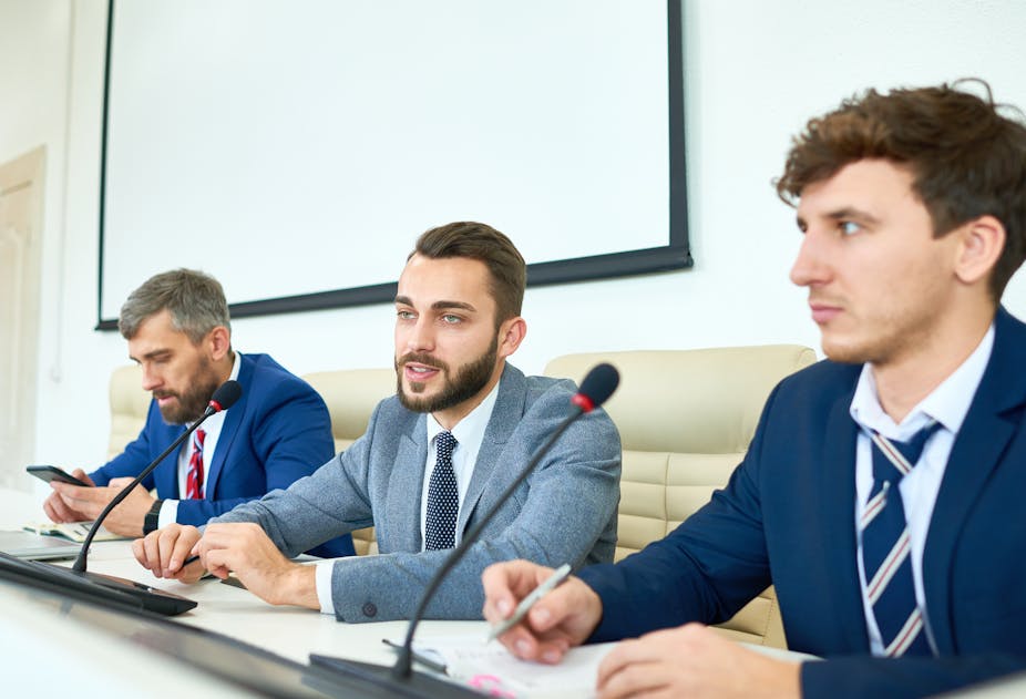 Three men in suits sat in front of microphones