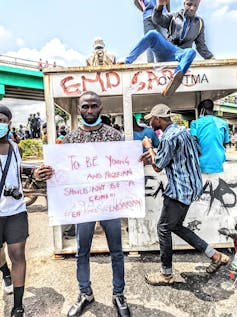 A young man with a beard displays a placard reading, 'To be young and Nigerian should not be a crime #ENDSARSNOW' as protesters move past in the background.