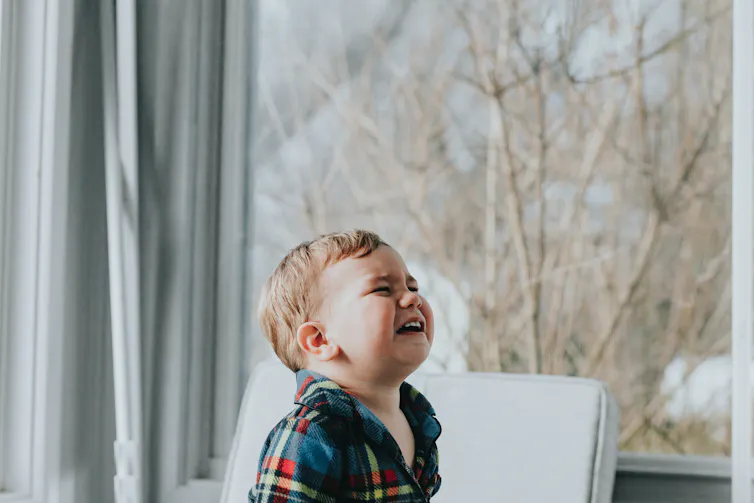 A baby boy in a plaid shirt throws his head back and cries.