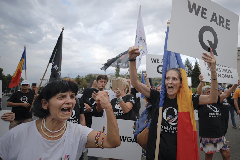 Angry women shaking their fists while one holds a sign in English that says We Are Q