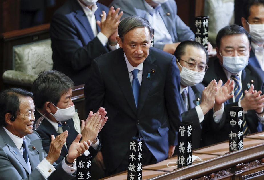 Yoshihide Suga stands up in parliament surrounded by seated men clapping.