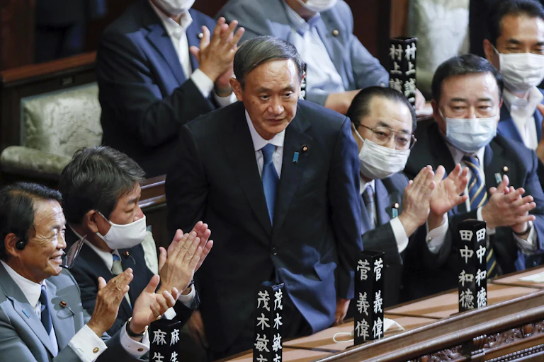 Yoshihide Suga stands up in parliament surrounded by seated men clapping.