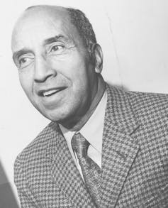 African American sports reporter Sam Lacy grins in a black-and-white portrait from 1960.