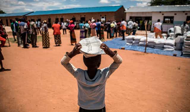 An African child balances a package on his head, with adults standing nearby alongside sacks of food.