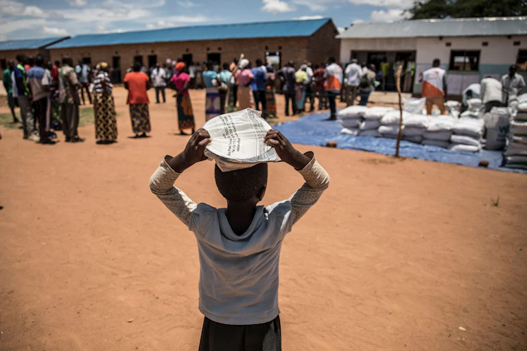An African child balances a package on his head, with adults standing nearby alongside sacks of food.