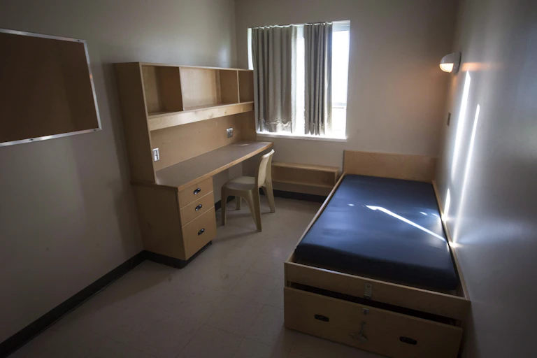 An empty cell with a cot, desk and bulletin board
