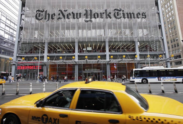 A glass building - with The New York Times written across it. In the foreground, a yellow cab.