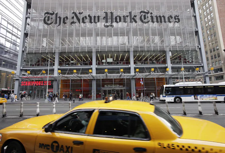 A glass building - with The New York Times written across it. In the foreground, a yellow cab.
