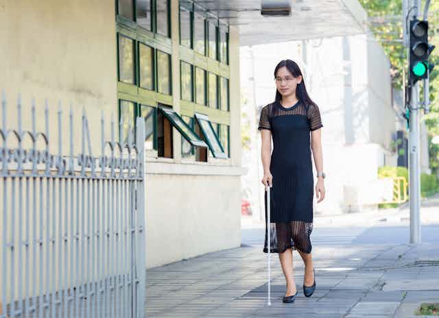 A young woman with impaired vision wearing a black dress, walking on the sidewalk with a white cane.