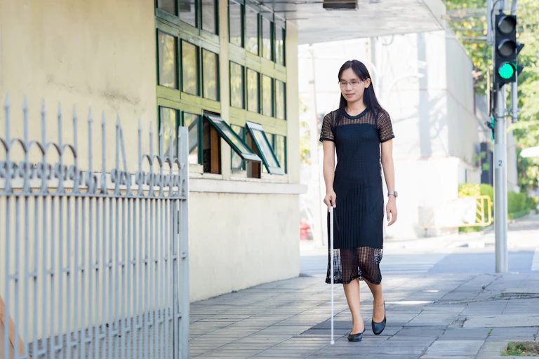 A young woman with impaired vision wearing a black dress, walking on the sidewalk with a white cane.