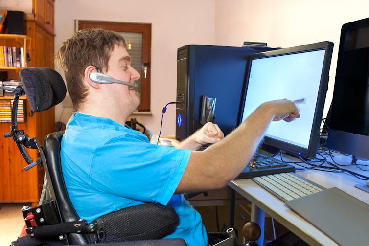 A young man with cerebral palsy in a wheelchair wearing a blue T-shirt and using a computer and a headset.