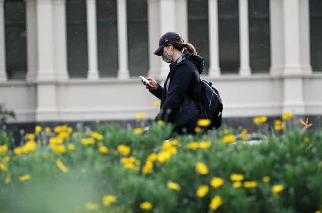 A lady, wearing a mask, looks at her phone will walking through Melbourne