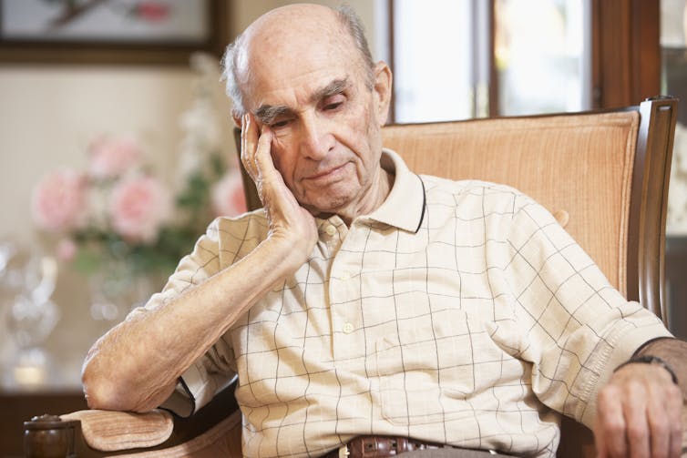 An elderly man, looking despondent, sits in a chair.