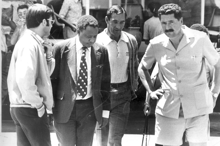 Percy Qoboza, wearing a dark suit and dark tie with white spots, surrounded by four white apartheid policemen.