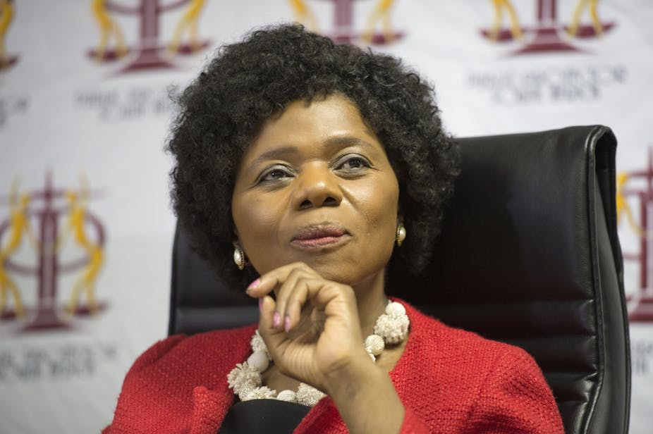Thuli Madonsela, wearing a black afro wig and red jacket in deep thought behind a desk.