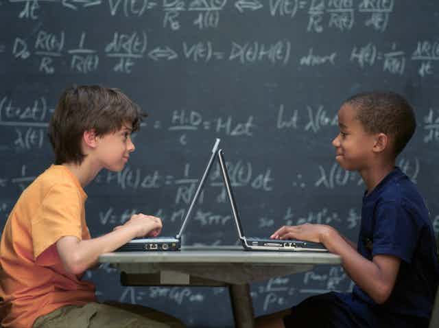Two boys sitting at desk with laptops in front of then and a blackboard in the background with maths equations.