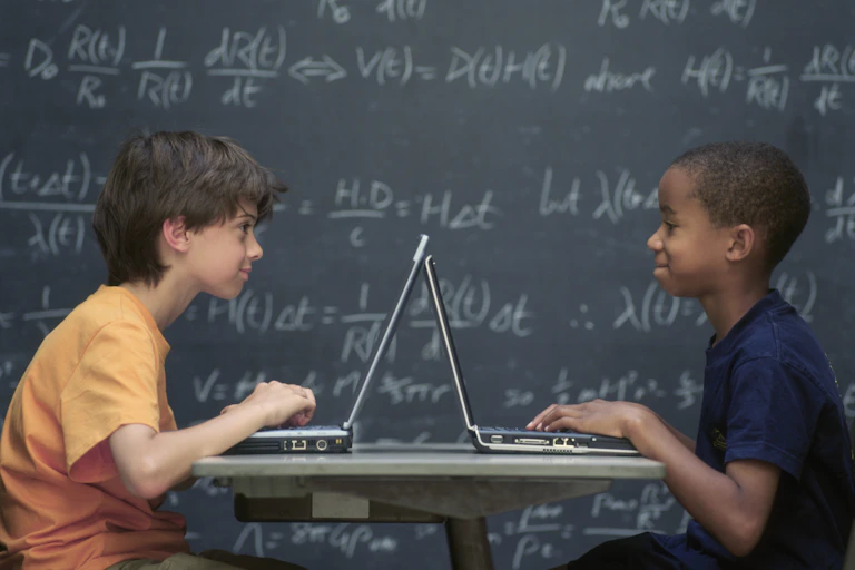 Two boys sitting at desk with laptops in front of then and a blackboard in the background with maths equations.
