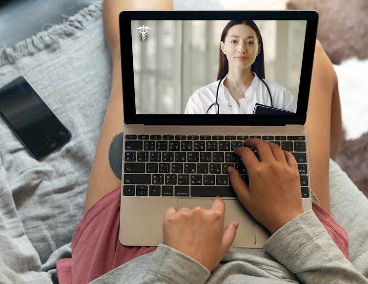 A woman in casual clothes on a sofa consulting with a doctor on her laptop.