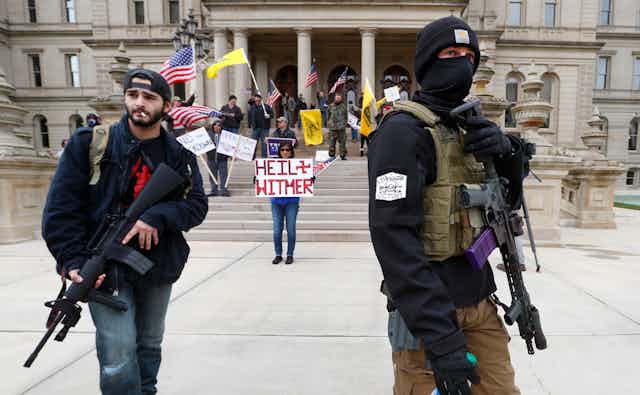 Two men stand in front of a government building carrying assault weapons while a woman carries a sign reading Heil Witmer, with Whitmer spelled incorrectly