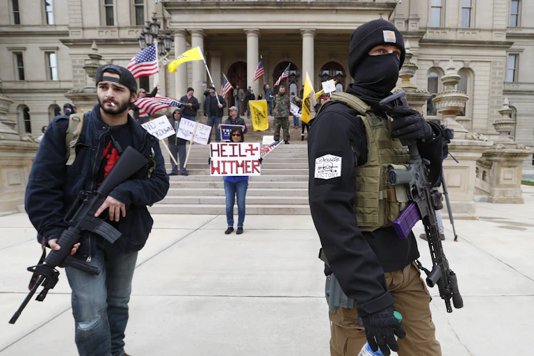Two men stand in front of a government building carrying assault weapons while a woman carries a sign reading Heil Witmer, with Whitmer spelled incorrectly