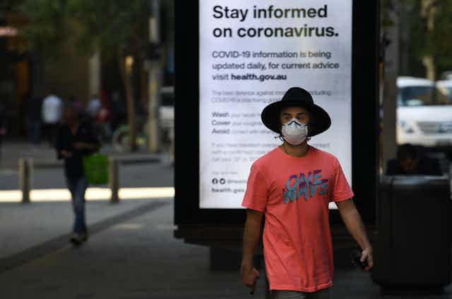 A man wearing a hat and facemask walks in front of a sign reading "Stay informed on coronavirus".