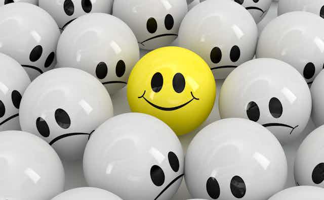 A yellow ball with a smiley face among several grey balls with frowny faces