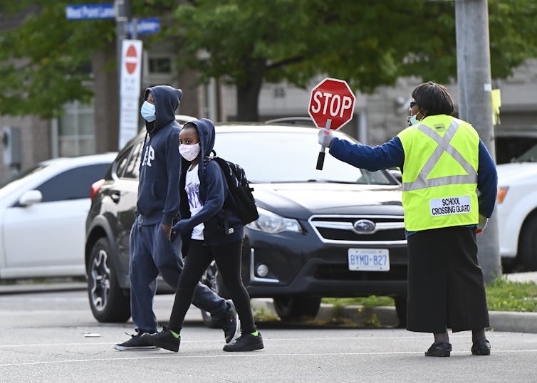 Two children cross the street; behind them a cross guard in a yellow vest holds up a stop sign.