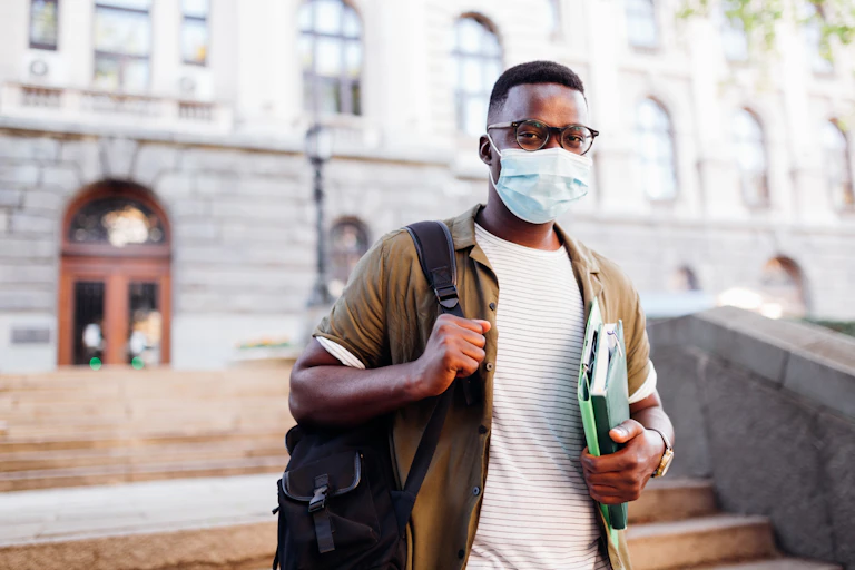 An African-American man wearing a mask walks down a flight of stairs while caring books.
