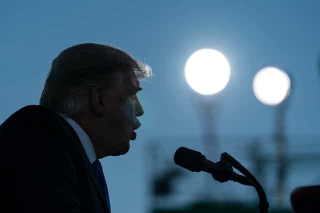 Donald Trump is seen in profile in the twilight with two shining lights in the background and microphone in front of him.