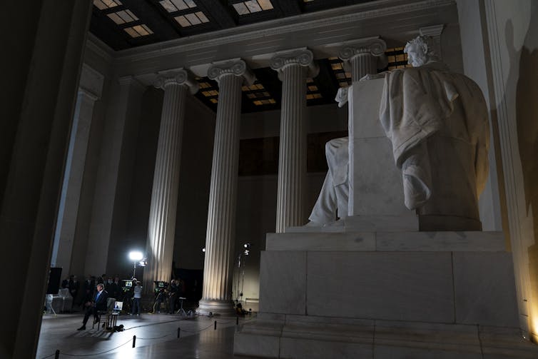 The Lincoln Memorial is in the foreground as TV lights illuminate a sitting Donald Trump