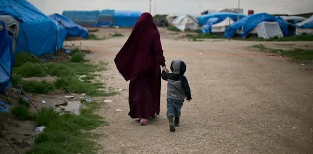 A woman wearing a burgundy hijab walks hand in hand down a dirt road with a young boy wearing a grey hodie.