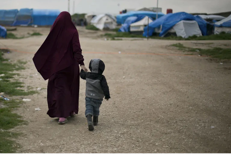A woman wearing a burgundy hijab walks hand in hand down a dirt road with a young boy wearing a grey hodie.