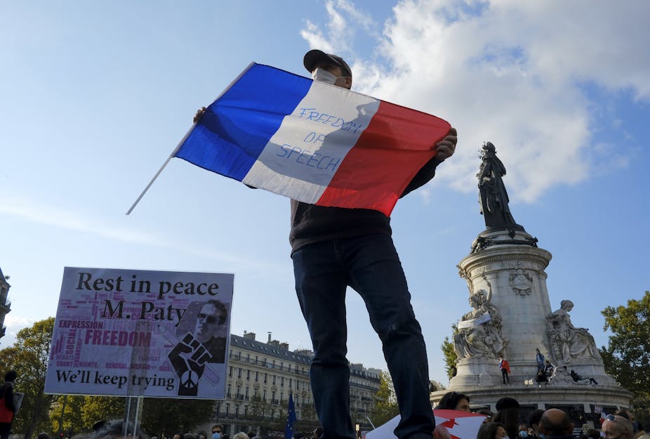 A man at a rally stands holding a French flag reading 'Freedom of Speech'