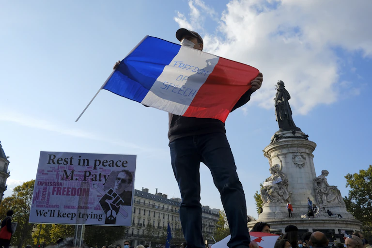 A man at a rally stands holding a French flag reading 'Freedom of Speech'