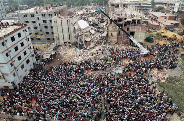 Hundreds of people are seen in an aerial shot in front of a collaposed multi-storey building.
