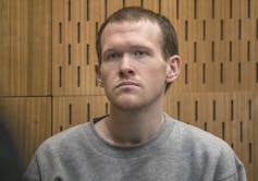 A short-haired white man in a grey sweatshirt sits in a courtroom dock.