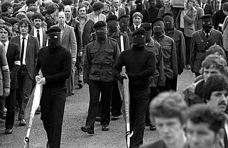 Masked men in black walk along a street carrying unfurled flags.