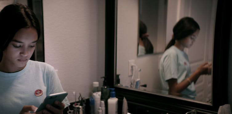 A young woman in a bathroom is engaged with her mobile phone, reflected in a mirror.