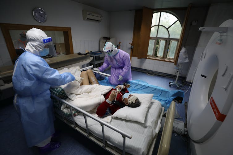 Man lies in hospital bed with two nurses next to him in full personal protective equipment.