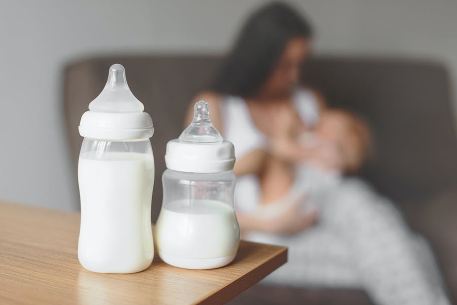 2 botol susu bayi dengan ibu menyusui di latar belakang. 