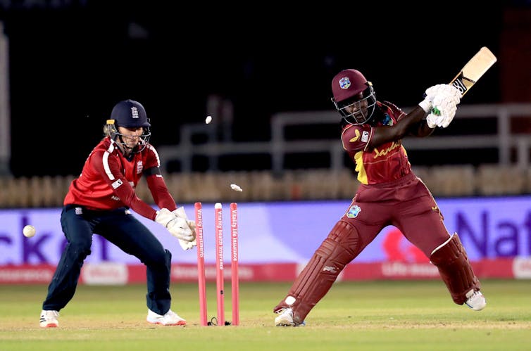 A woman cricket player swings her bat while another hits the stumps.